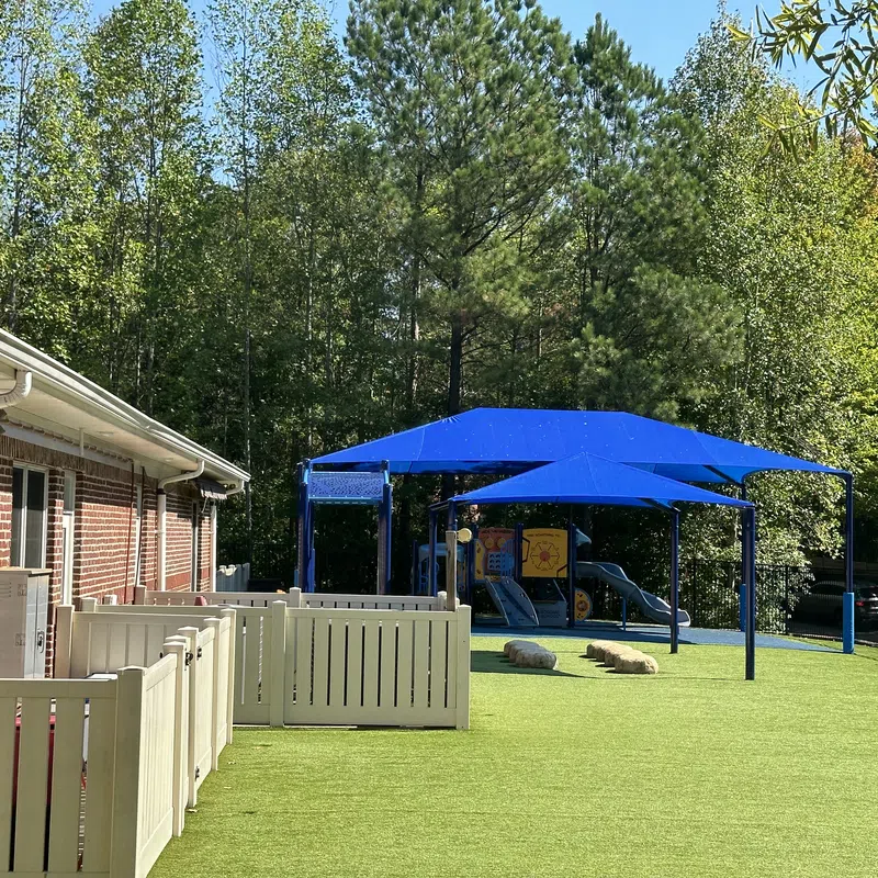 Outdoor playground with blue shade sails, artificial grass, play structures, and fenced area next to brick building.
