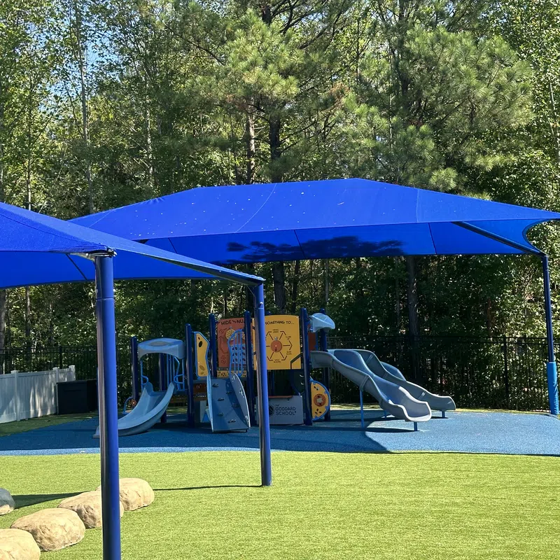 Outdoor playground with blue slides, climbing structures, and blue shade canopies surrounded by greenery.
