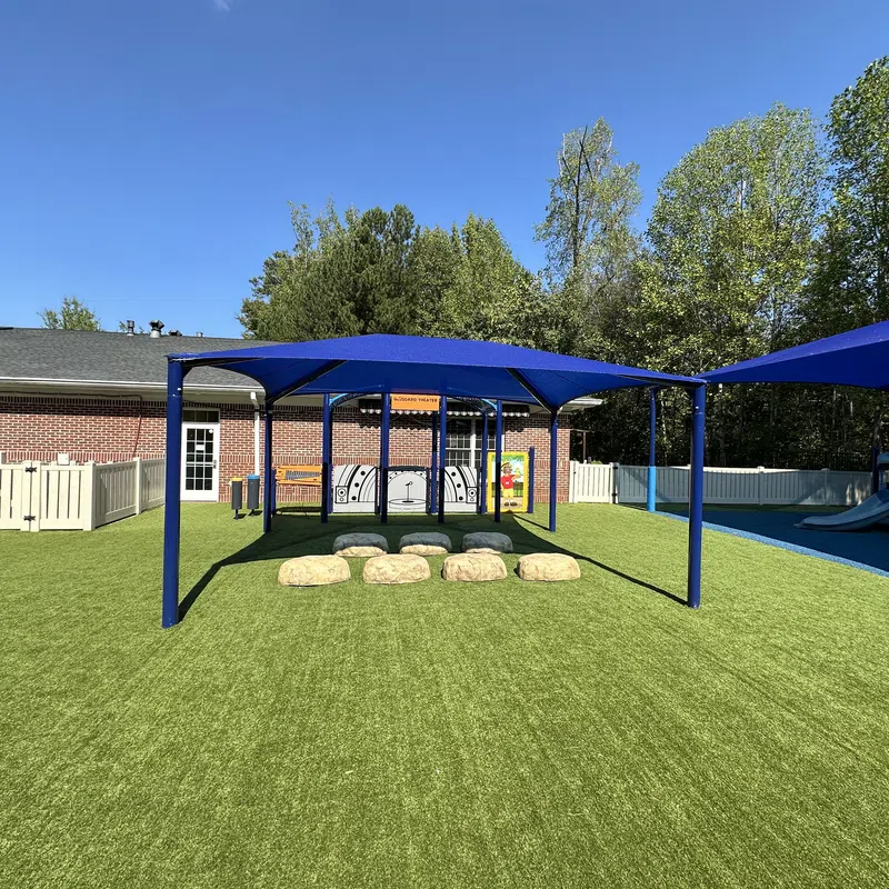 Playground area with artificial grass, blue shaded canopies, stepping stones, and a brick building in background