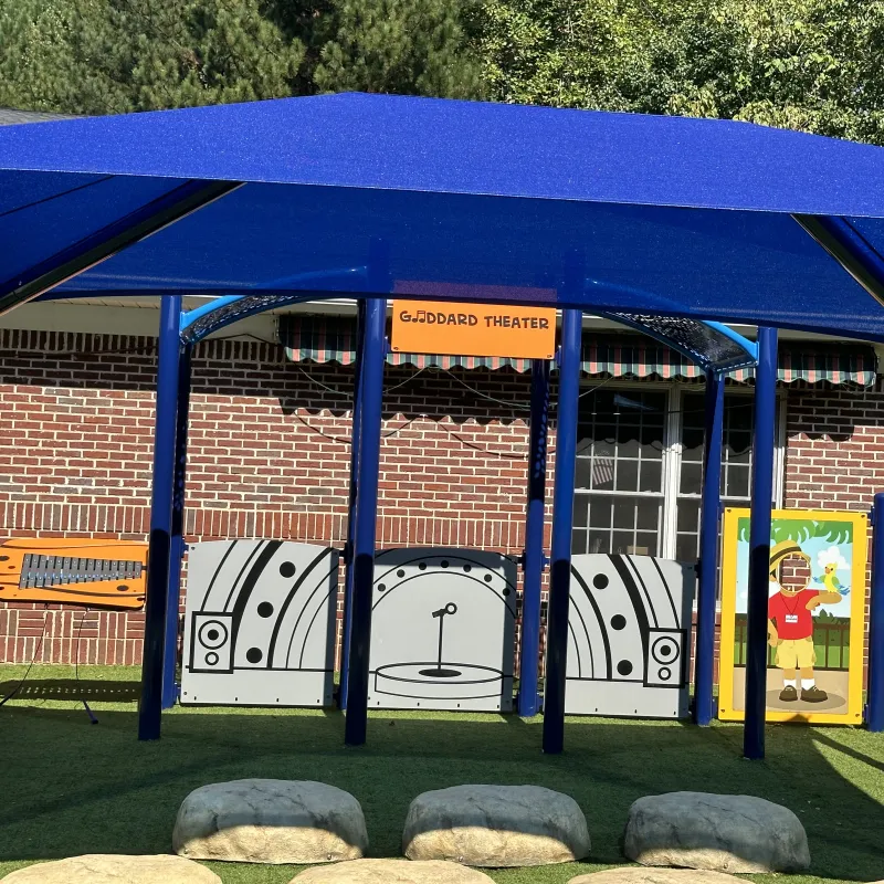 Outdoor children's play theater area with blue canopy, stage backdrop, and artificial grass seating resembling rocks.