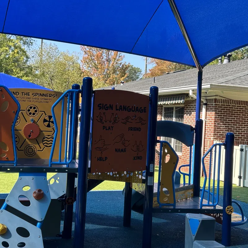 Children's playground with sign language learning panel and colorful climbing structures under a blue shade canopy.