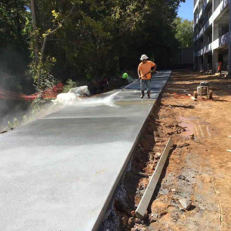 Construction worker spraying water on freshly poured concrete sidewalk beside building and trees on sunny day