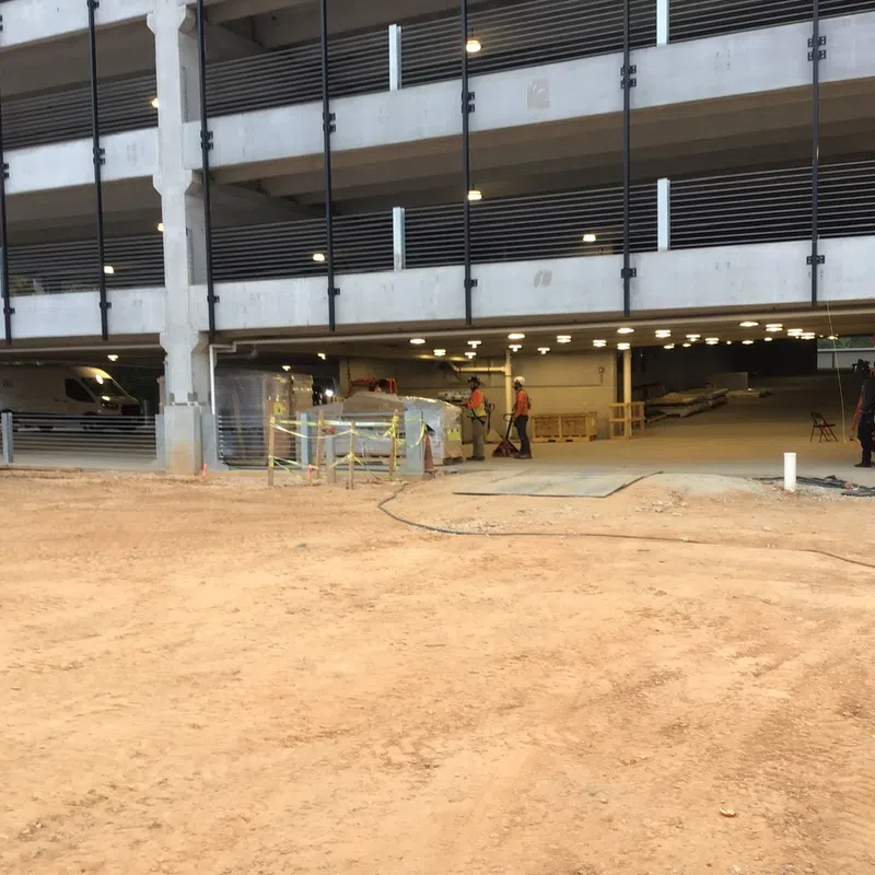 Construction site outside a multi-level concrete parking garage with workers and equipment visible.