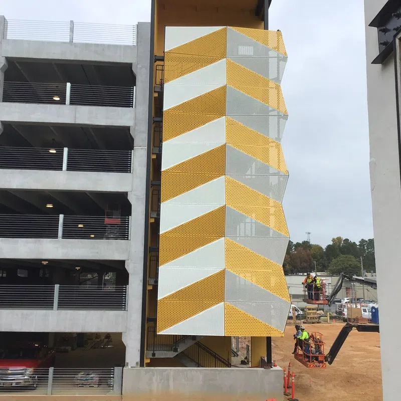 Construction workers install yellow and white chevron-patterned metal panels on a stairwell beside a concrete parking garage.