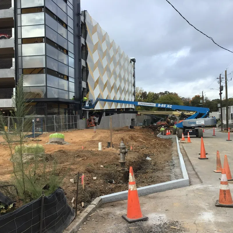Construction site with a modern building facade, orange safety cones, and workers operating a boom lift under cloudy sky.