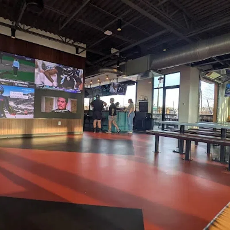 Modern sports bar with large screens showing games, industrial ceiling, wooden benches, and people near the bar counter.