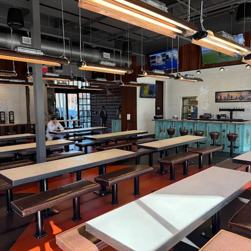 Modern industrial-style cafeteria with long tables, bench seating, hanging lights, and a bar area in the background