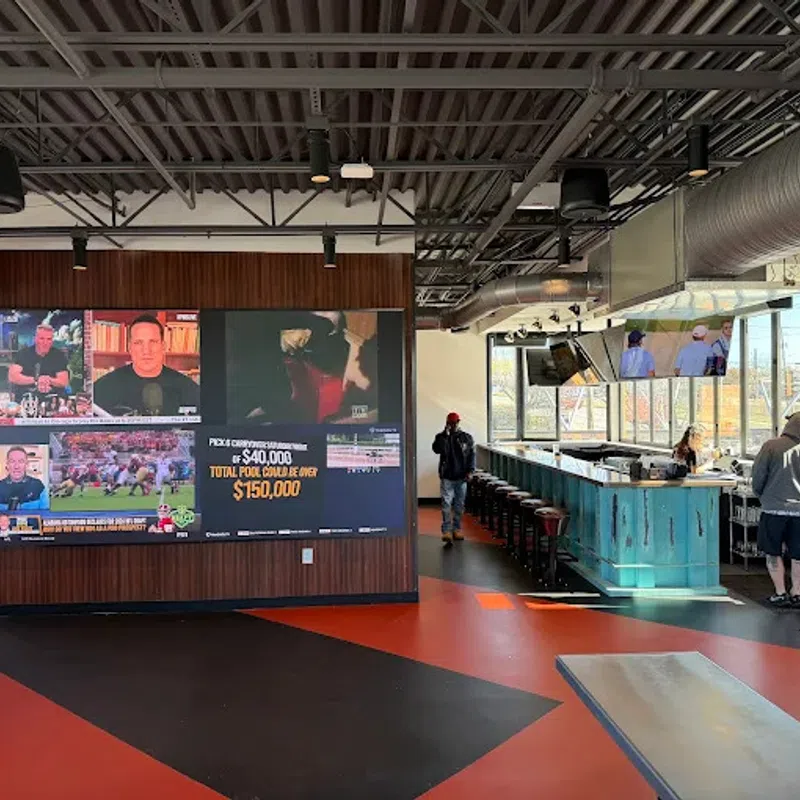 Modern sports bar interior with large TV wall, blue bar counter, industrial ceiling, and customers inside.