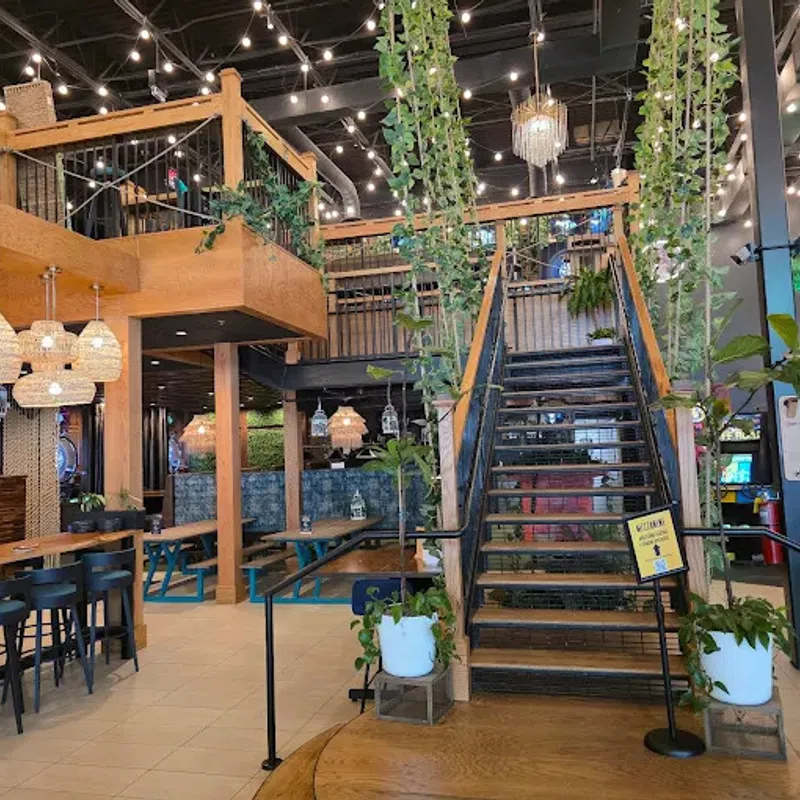 Modern indoor restaurant with wooden tables, hanging lights, plants, staircase, and arcade games in the background