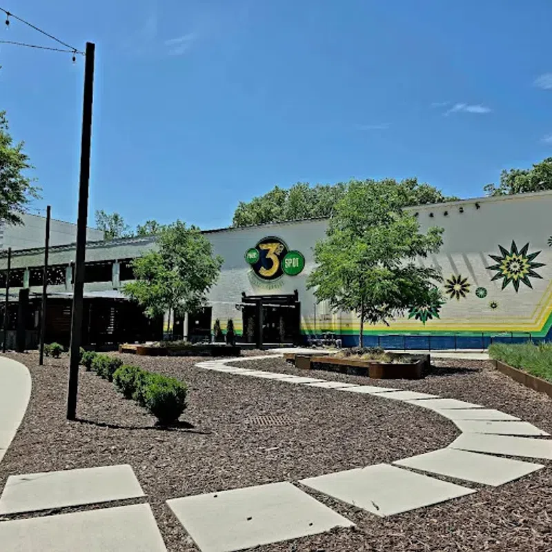 Outdoor view of The 3 Spot building with curved sidewalk, trees, and decorative mural under blue sky.