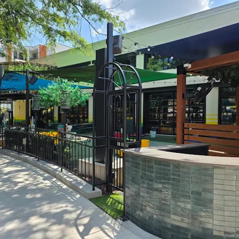 Outdoor patio area with shaded seating, black metal fencing, tiled counter, and blue umbrella on a sunny day.