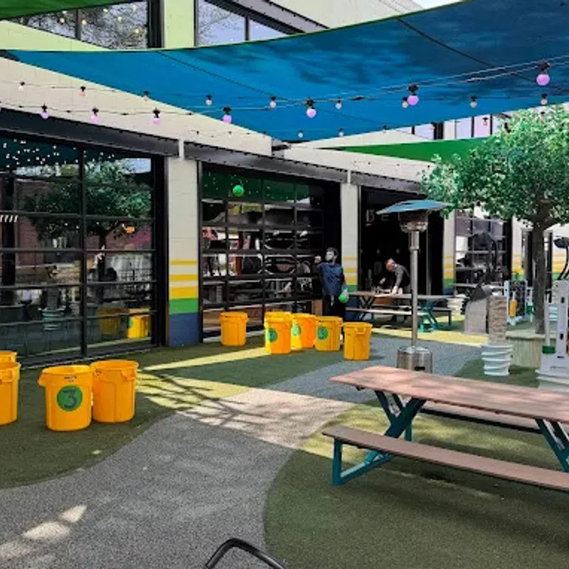 Outdoor seating area with picnic tables, yellow recycling bins, shade sails, and string lights in a commercial space.