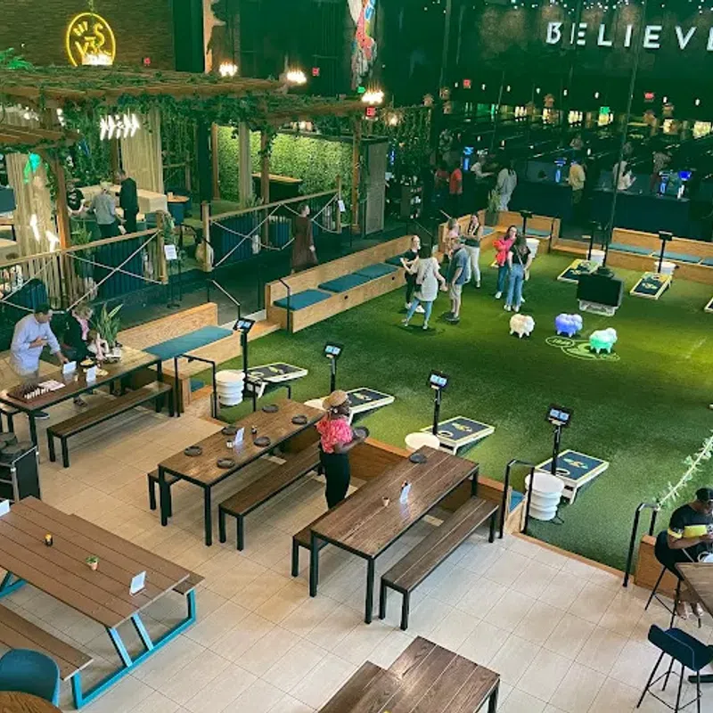 Indoor recreational space with people playing cornhole, sitting at wooden tables, and green turf floors under plants.