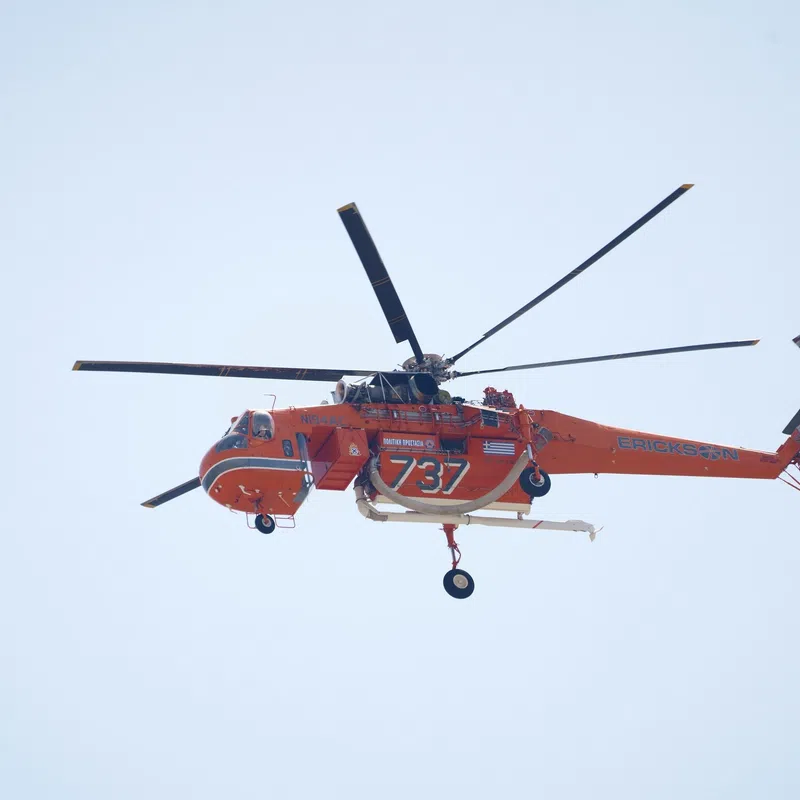 Orange Sikorsky S-64 Skycrane helicopter flying against clear sky with firefighting equipment visible.