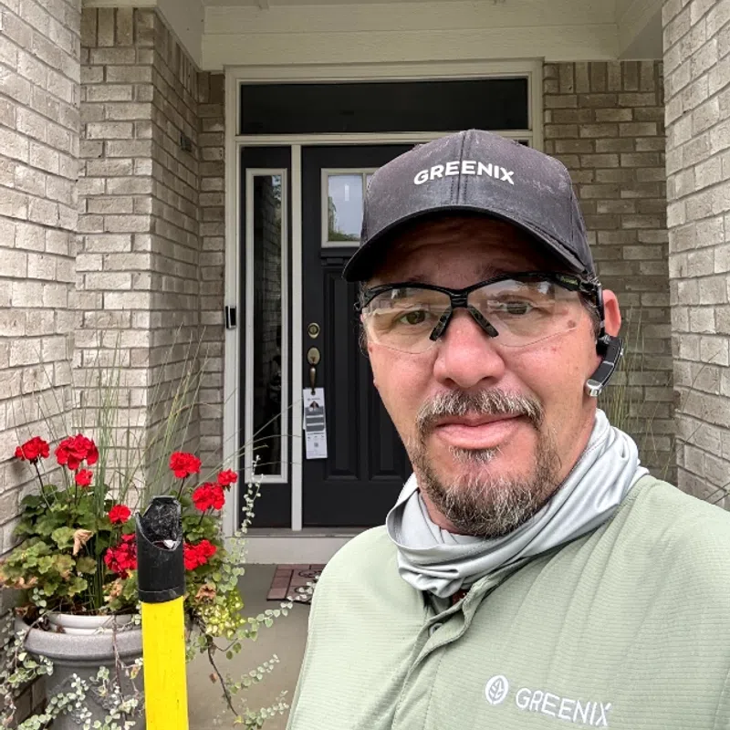 Man wearing Greenix cap and shirt standing in front of a brick house entrance with red flowers and black door.
