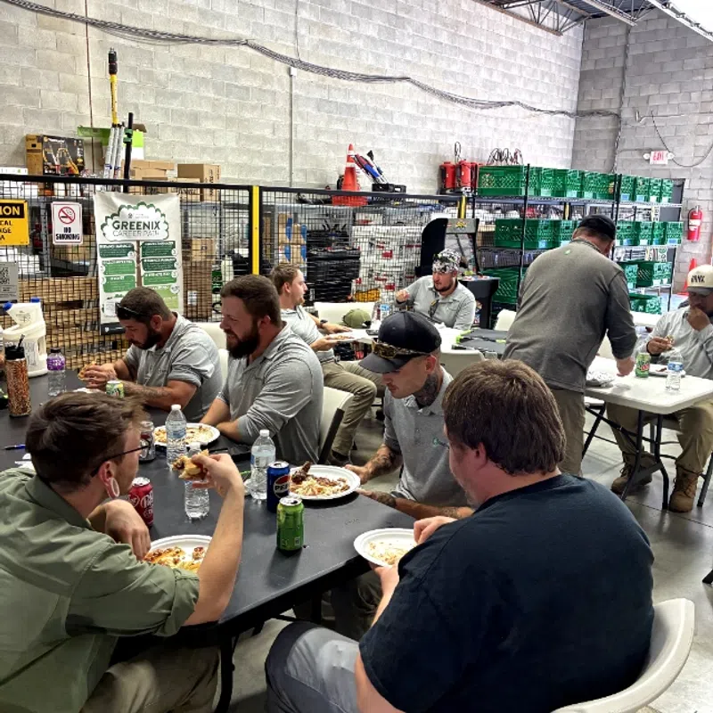 Workers eating lunch together at tables inside a warehouse with stacked boxes and industrial shelves.