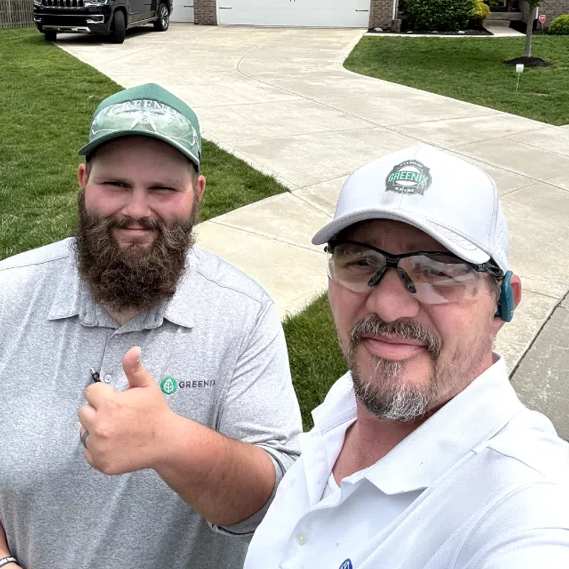 Two men in Greenix uniforms smiling with thumbs up in front of a clean driveway and house.