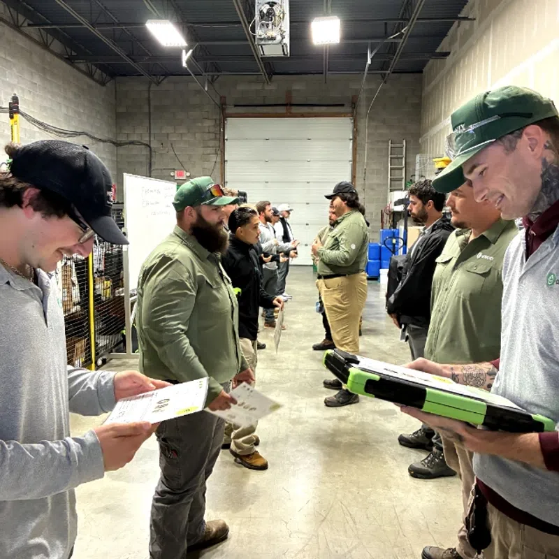 Workers in casual attire stand in two lines facing each other inside a warehouse holding papers and tablets.