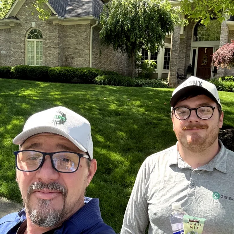 Two men wearing glasses and branded caps stand outside a brick house with green lawn and trees on a sunny day.