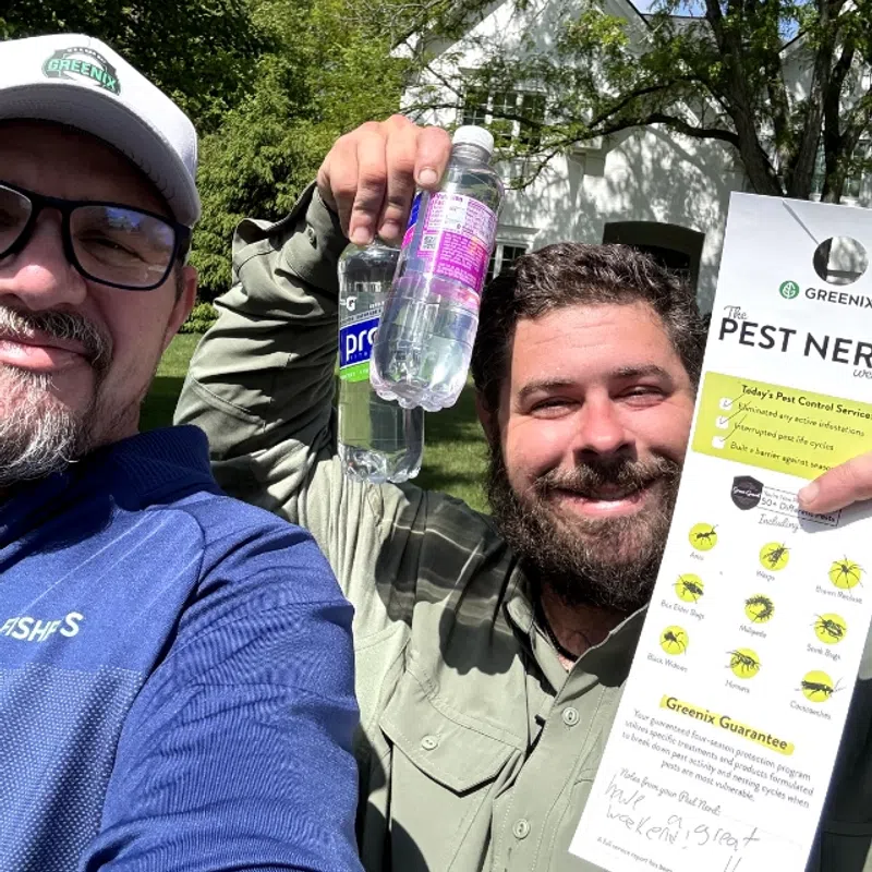 Two men outdoors smiling, holding water bottles and a pest control flyer in front of a white house and trees.