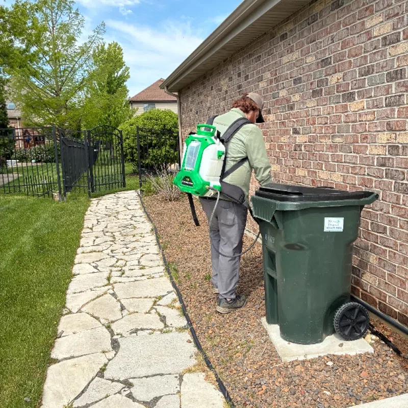 Person wearing green backpack sprayer disinfecting green outdoor trash bin beside brick house on sunny day.