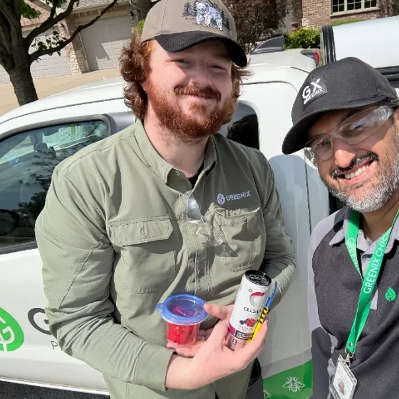 Two pest control workers smiling in front of a Greenix branded vehicle, holding a snack and a drink.