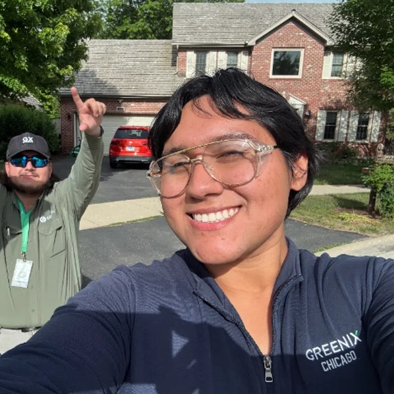 Two people smiling outdoors in a residential neighborhood with trees and a brick house in the background.