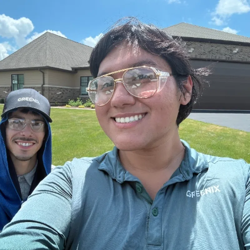 Two smiling men in Greenix uniforms outdoors on a sunny day with houses and green lawn in the background