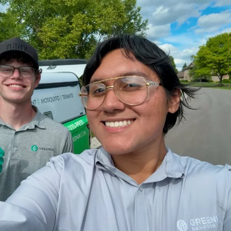 Two pest control workers smiling in uniform by a company van on a sunny suburban street.