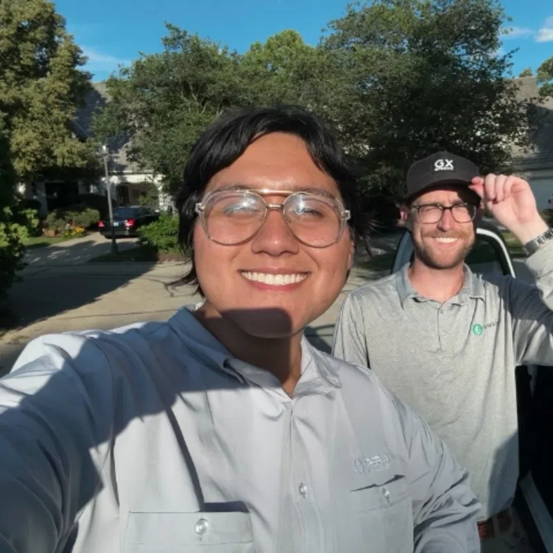 Two men smiling outdoors near a white vehicle in a residential neighborhood on a sunny day.