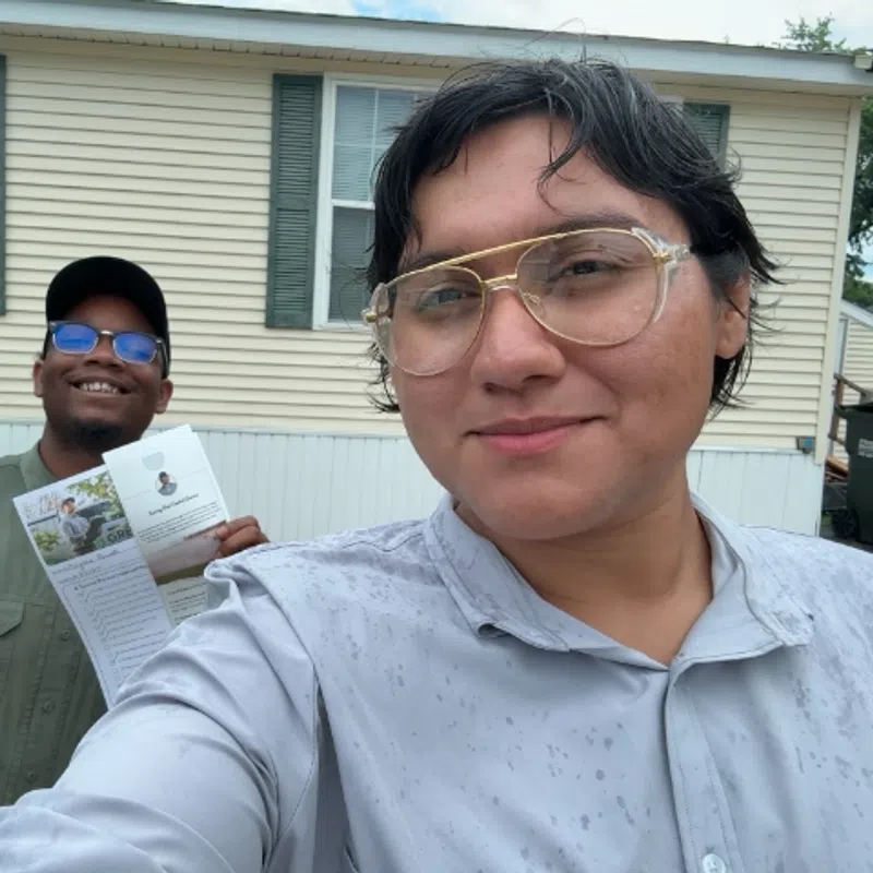 Two smiling people taking a selfie outdoors in front of a house, one holding a flyer or brochure.