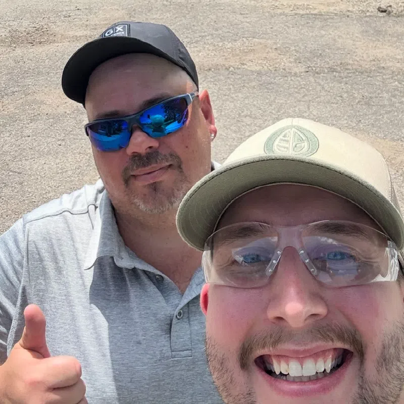 Two men wearing hats and sunglasses smiling outdoors on a sunny day with green trees and blue sky behind them.