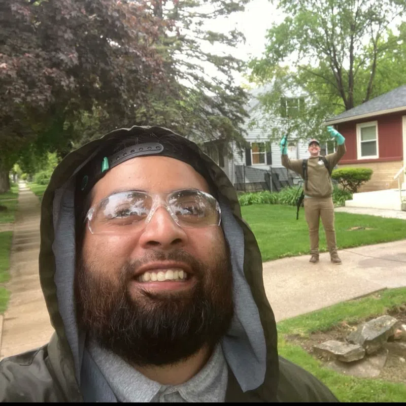 Two men outdoors on a residential sidewalk, one smiling close to camera, the other waving in the background near houses.