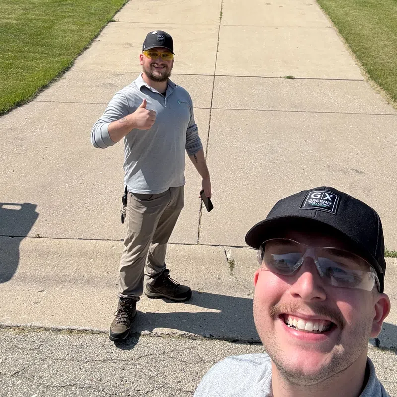 Two men in work attire smiling and posing in front of a suburban house driveway on a sunny day.