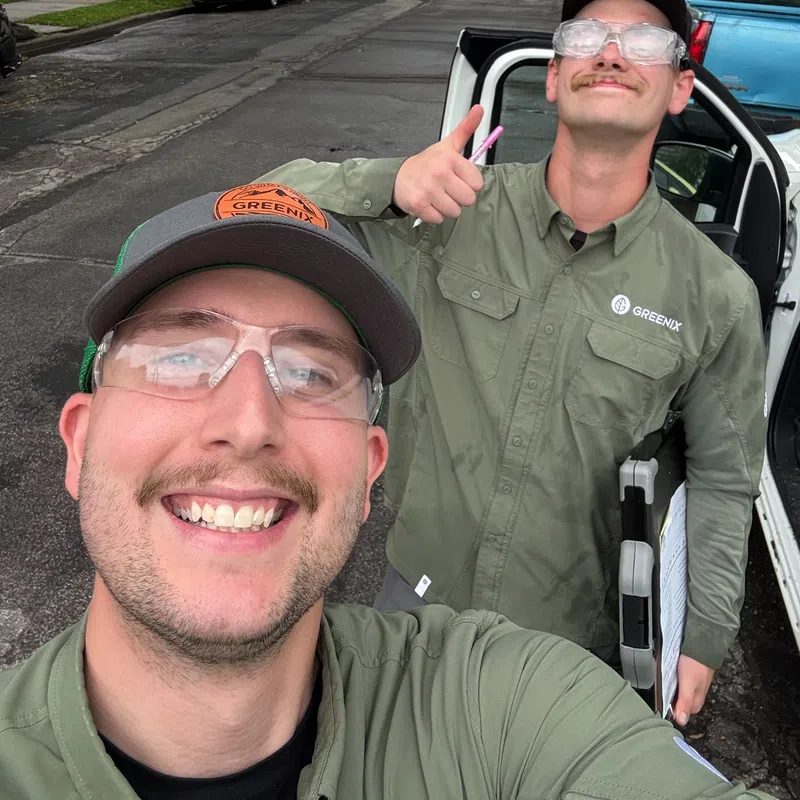 Two smiling landscapers in green uniforms and safety glasses pose for a selfie on a residential street.