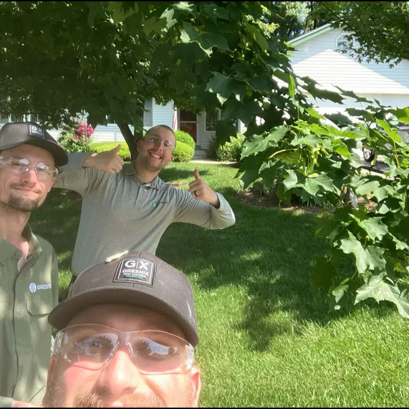 Three landscapers wearing safety glasses and caps posing outdoors in front of a house with green lawn and trees.