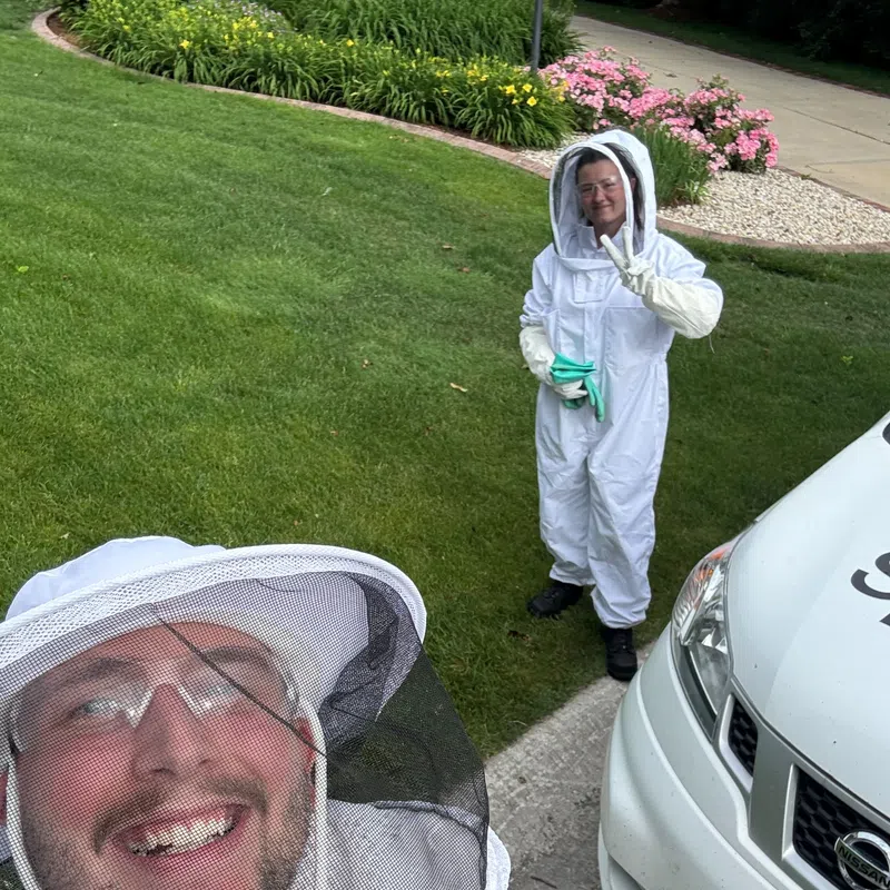 Two people in white beekeeping suits smiling near a white van and green lawn with flowers.