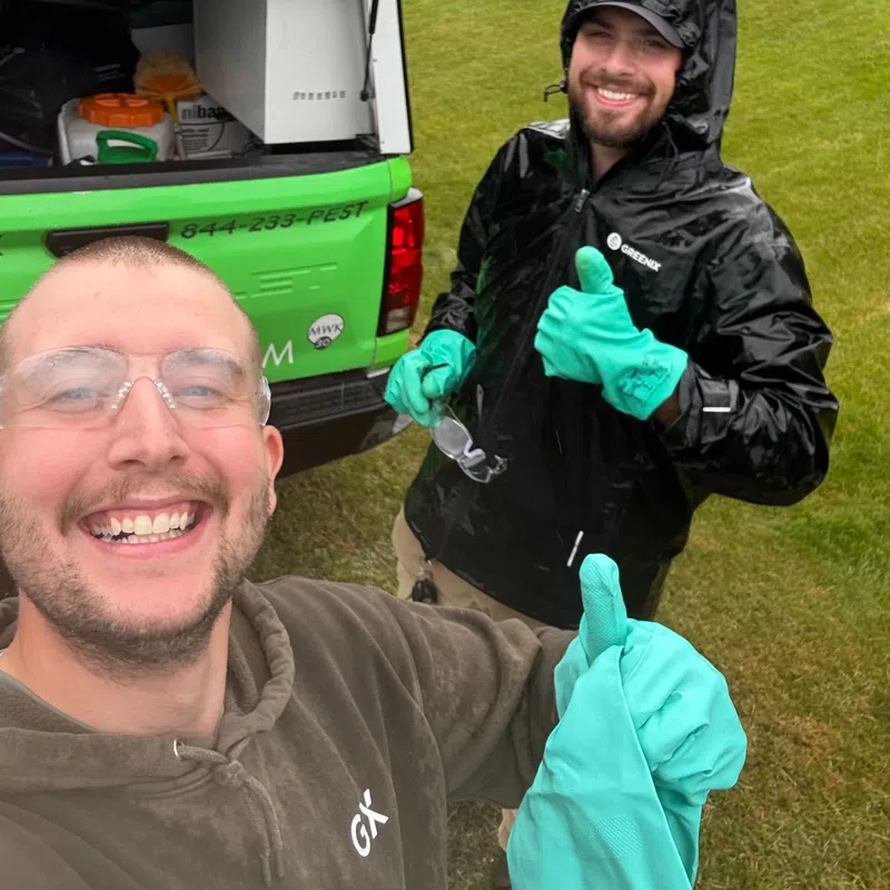 Two pest control workers in protective gloves giving thumbs up next to a green truck on a grassy lawn.