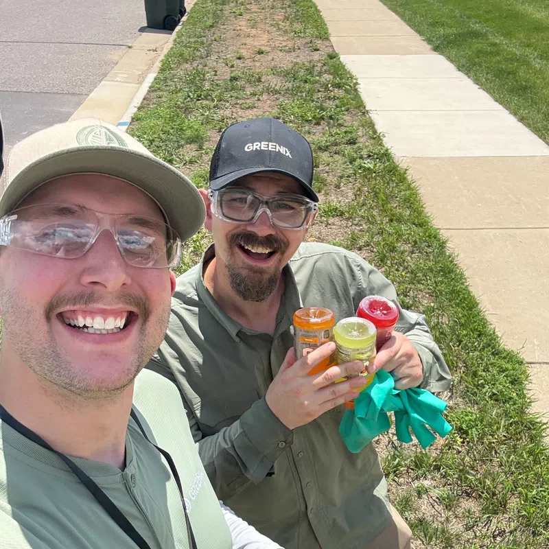 Two men in work uniforms smiling outdoors, one holding containers and gloves, standing on grass by a sidewalk.