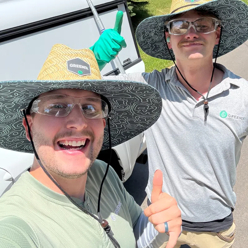 Two workers in Greenix hats and safety glasses giving thumbs up outside on a sunny day near a trailer.