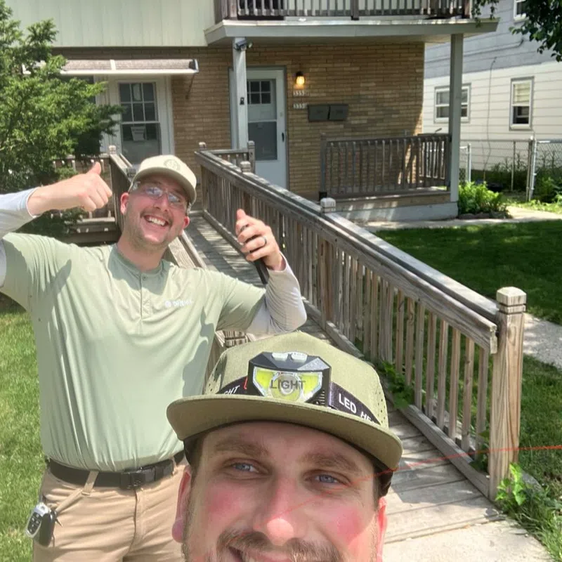 Two smiling men outdoors, one giving thumbs up, standing near a wooden ramp and green lawn in front of a house.