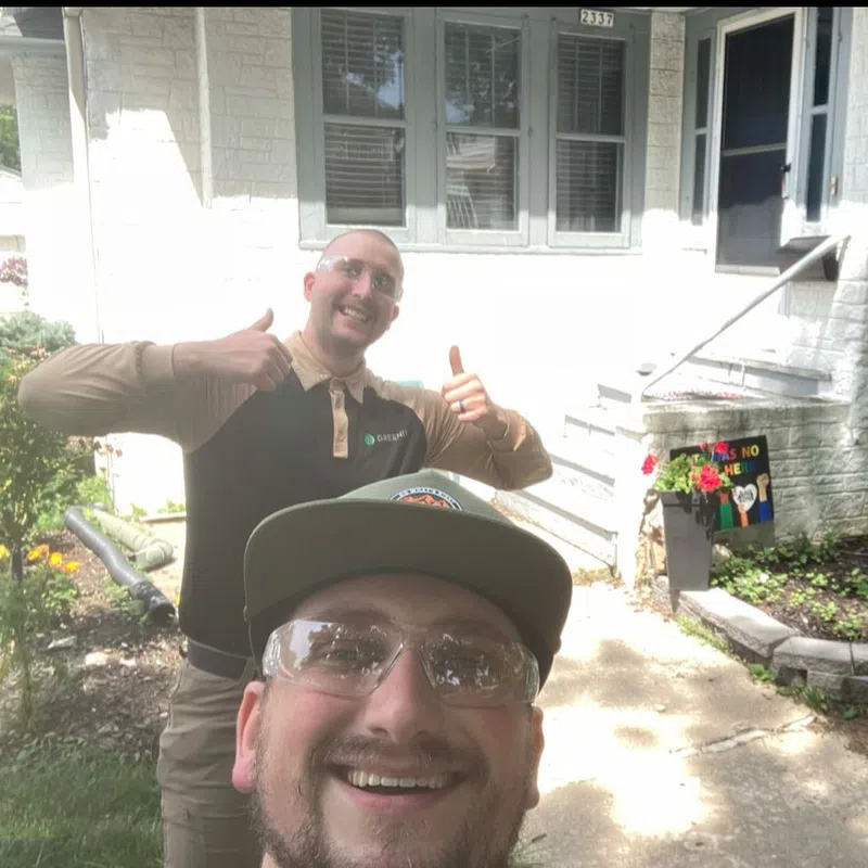 Two men smiling and giving thumbs up outside a white house with garden and steps.