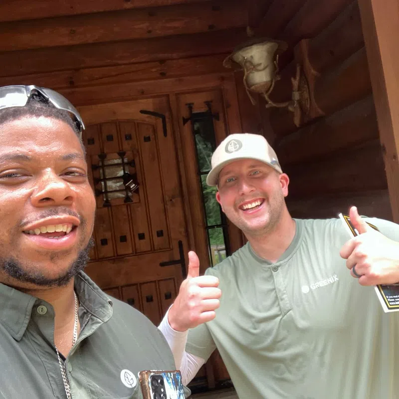 Two men smiling and giving thumbs up outside a wooden cabin entrance with rustic decor.