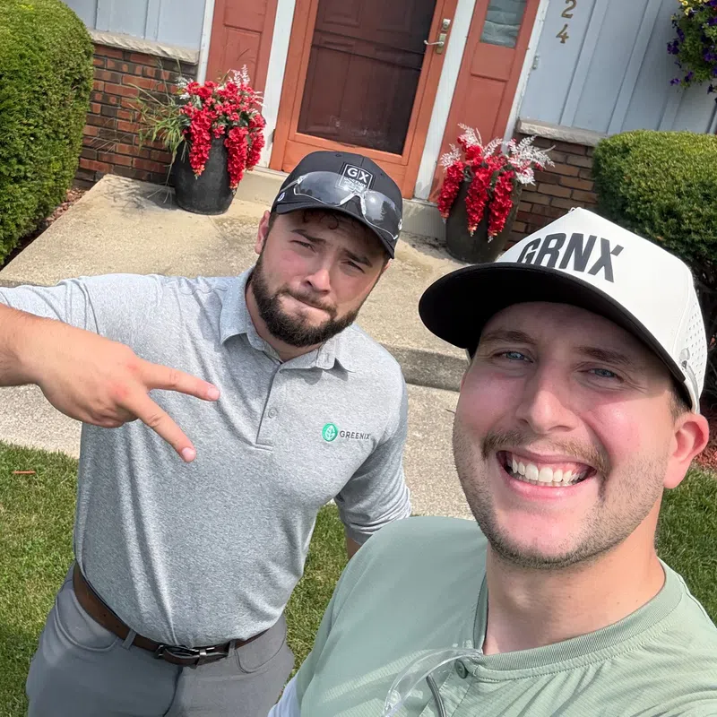 Two men smiling outdoors in front of a house entrance with red flowers and green bushes on a sunny day.