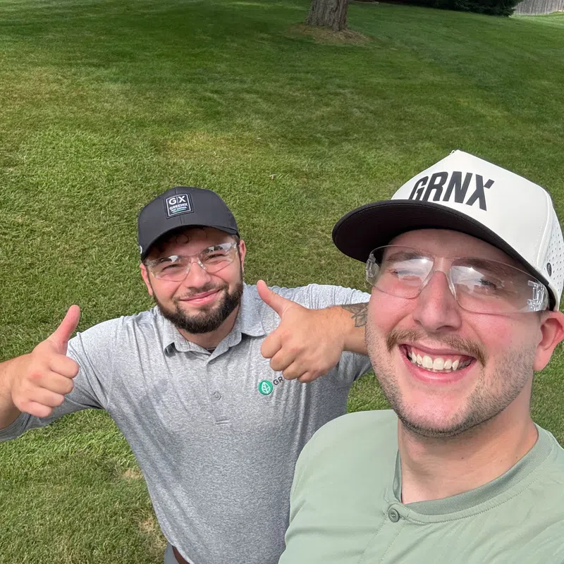 Two smiling men wearing caps and safety glasses give thumbs up on a well-maintained green lawn in front of a house.