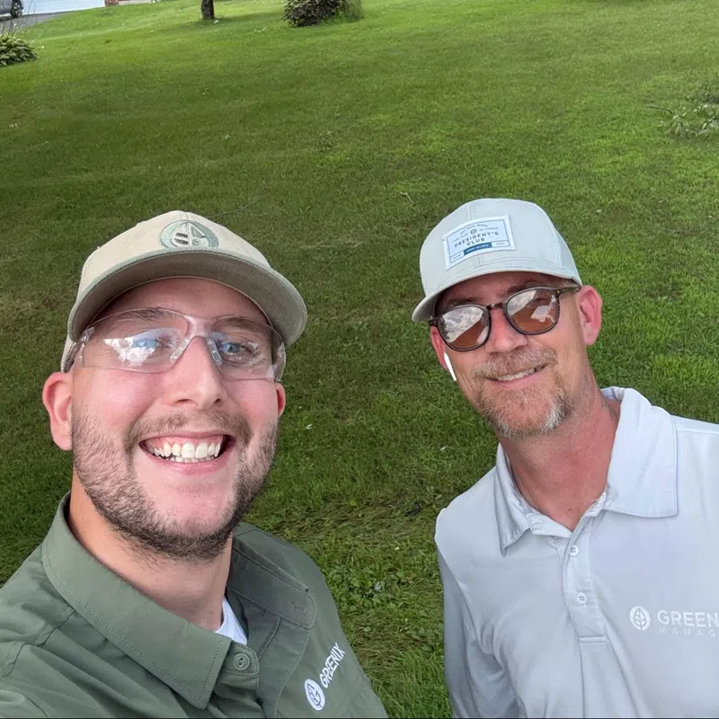 Two men smiling in lawn care uniforms standing on a green grassy yard with trees and houses in the background.