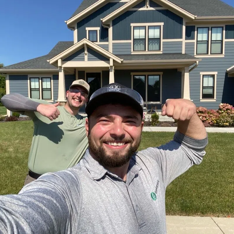 Two men smiling and flexing muscles in front of a large blue house on a sunny day