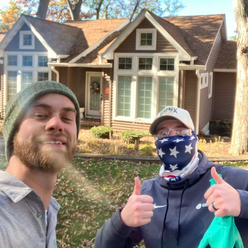 Two men giving thumbs up in front of a brown house with large windows on a sunny autumn day.