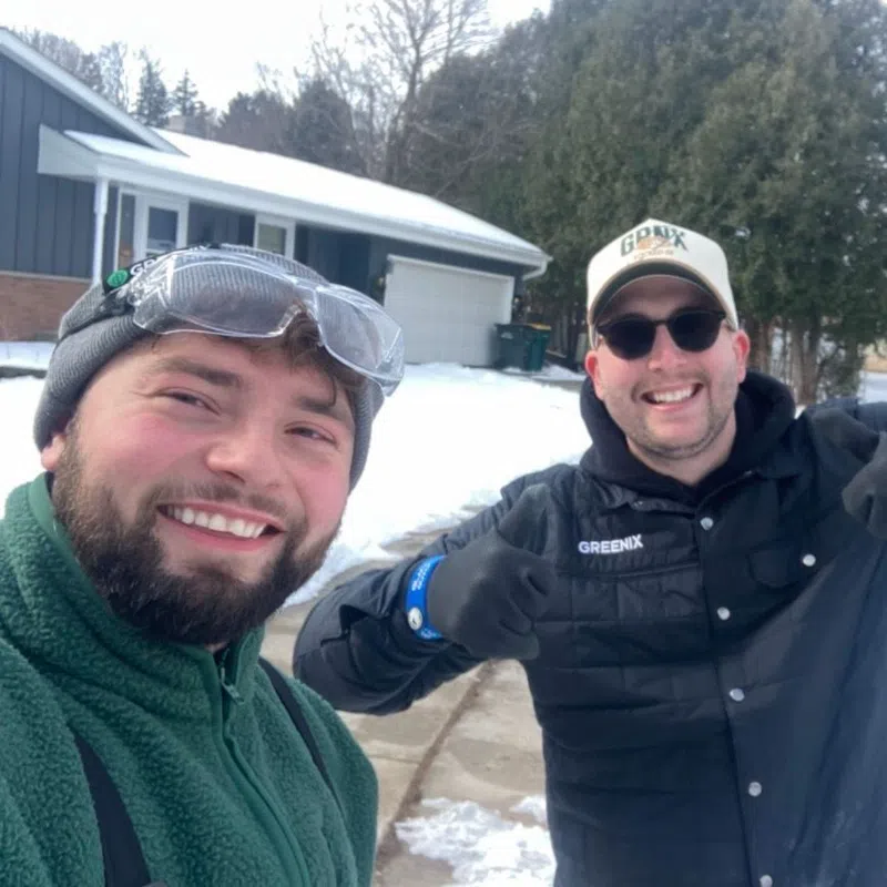 Two men smiling outdoors in winter gear with snow and houses in the background on a cold day.