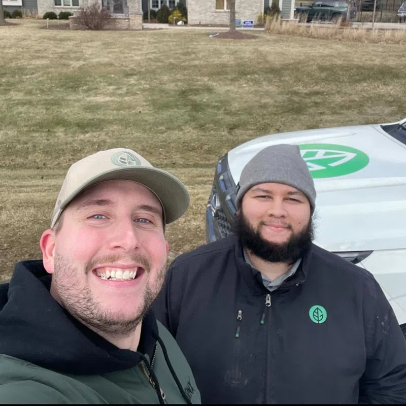 Two smiling men in outdoor jackets and hats stand in front of a large suburban house and a white vehicle with green leaf logo.